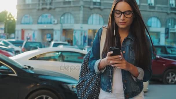Hermosa mujer joven caminando por la calle y escuchando música en auriculares desde el teléfono inteligente. Ciudad calle fondo — Vídeos de Stock