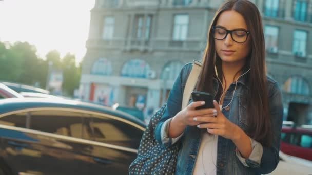 きれいな若い女性、街を歩くと、スマート フォンのイヤホンから音楽を聴いてします。街の背景。内気な少女 — ストック動画