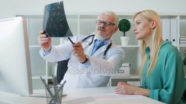 Onun offise oturan, kadın hasta x-ray görüntü için gösterilen ve hastaya konuşurken orta aded beyaz kıdemli doktor adam. Kapalı.