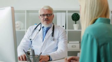 Beyaz genç bayanla konuşmak ve bir şey hastaya bilgisayarda gösterilen gözlüklü doktor beyaz orta yaşlı.