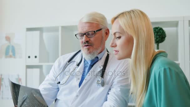 Grand médecin caucasien assis dans son bureau, parlant à une patiente, montrant une image radiographique du patient. Intérieur . — Video