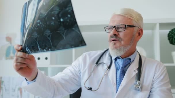 Retrato de médico caucásico de mediana edad en gafas con barba sentado en su consultorio y mirando imagen roentgen. De interior . — Vídeos de Stock