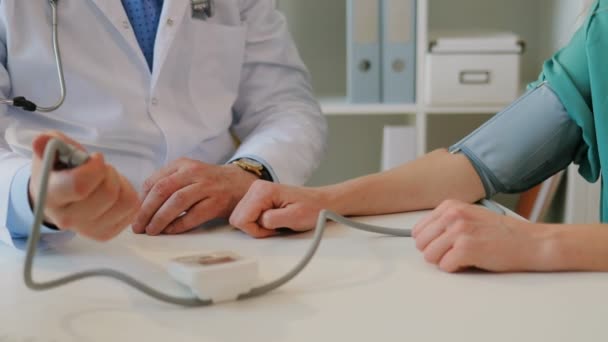 Кавказский врач в белом халате сидит в своем кабинете и измеряет артериальное давление кавказского пациента. Закрыть . — стоковое видео