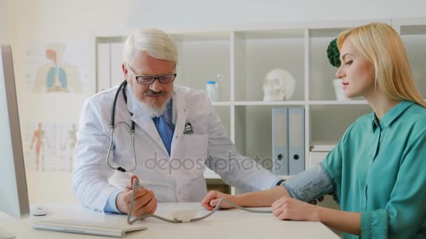 Médecin caucasien d'âge moyen en robe blanche avec des lunettes mesurant la pression artérielle caucasienne jeune patiente. Intérieur . — Video