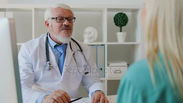 Caucásico de mediana edad médico varón leyendo algo de hoja de papel a paciente femenino, sonriendo, sentado en el escritorio en la oficina . — Vídeos de Stock