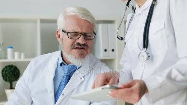 Beyaz orta yaş arası erkek doktor ofisinde oturan ve tablet kullanma ve bir şey hakkında konuşuyor ofis ayakta genç kadın doktor. Kapalı.