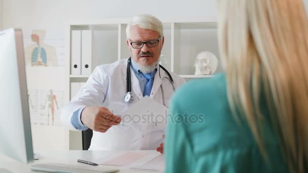 Kaukasische ernstige zijaanzicht leeftijd doctor in de witte robe met glazen en baard zitten in zijn kantoor iets met vel papier uit te leggen. Indoor. — Stockvideo