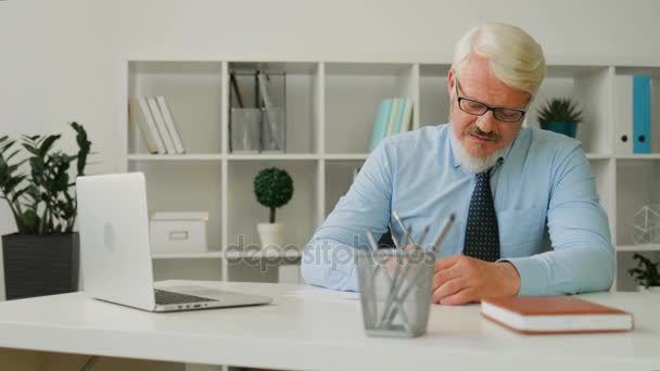 Volwassen Kaukasische man met bril in blauw overhemd en stropdas zitten op het Bureau op kantoor, het schrijven van iets op een vel papier. Indoor. — Stockvideo