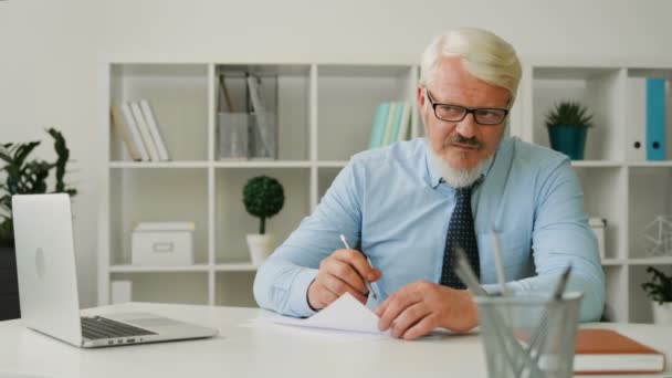 Белый мужчина средних лет в очках в голубой рубашке и галстуке сидит в офисе. Офицер что-то пишет на листе бумаги, сгибает и бросает бумагу, ломает карандаш в помещении . — стоковое видео