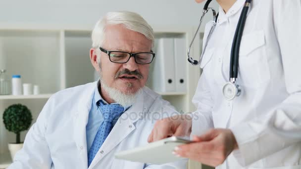 Médico de mediana edad caucásico sentado en su consultorio y joven médico de pie en el consultorio usando tableta y hablando de algo. De interior . — Vídeo de stock