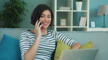 Dizüstü bilgisayar kullanarak ve oturma odasında kanepede otururken cep telefonuyla konuşurken güzel ve genç birbayan. Mutlu kadın konuşmayı