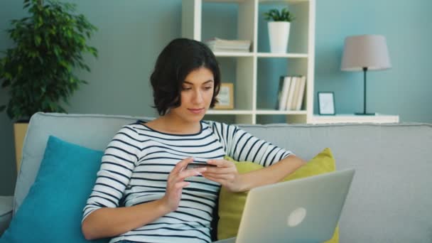 Aantrekkelijke vrouw met creditcard voor online winkelen op haar laptop zittend op de Bank in de woonkamer — Stockvideo