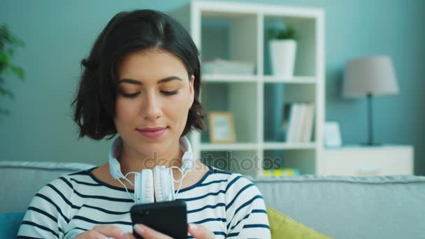魅力的な女性は、自宅でソファに座ってスマート フォンを使用してヘッドフォンを身に着けています。肖像画のショット — ストック動画