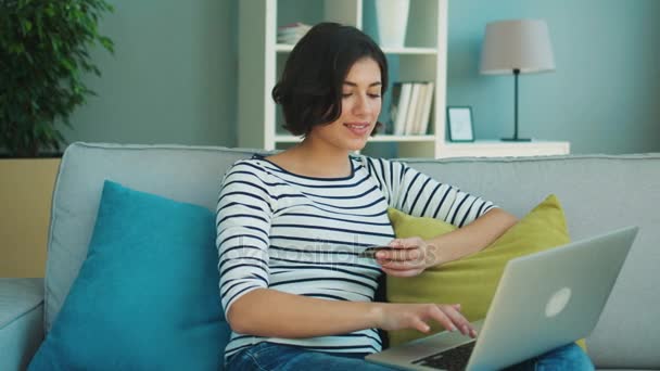Молодая брюнетка успешно зарегистрирована с помощью кредитной карты. Женщина делает онлайн покупки на ноутбуке, заявляя на диване . — стоковое видео