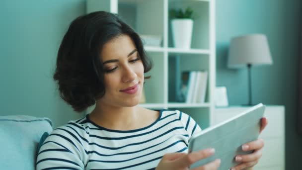 Mulher morena bonita sentada no sofá com computador tablet. Uma mulher feliz a sorrir. Cale-se — Vídeo de Stock