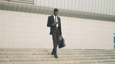 Merdivenlerden aşağı yürüme çekici siyah Afrika kökenli Amerikalı iş adamı adımları, akıllı telefon kullanarak. SMS okuma, internet'te sörf ve colegues için sallayarak adam. Adam eve işten sonra