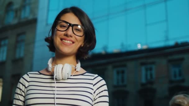 Retrato og hermosa joven con auriculares y gafas mirando directamente a la cámara y sonriendo. Ciudad calle, al aire libre — Vídeos de Stock