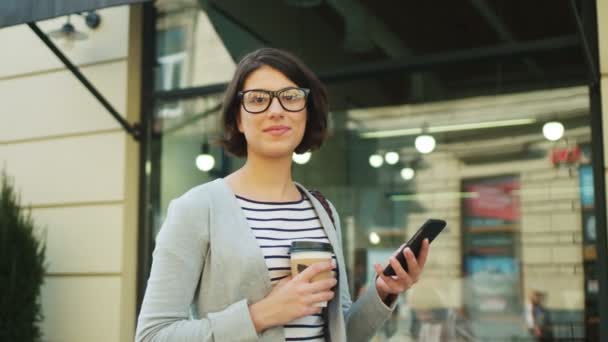 Portrait shot of woman using smart phone while standing ouside near cafe. Woman looking at the camera and smiling — Stock Video