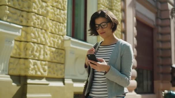 Mujer atractiva caminando por la calle de la ciudad con su teléfono inteligente y sonriendo a la cámara. De cerca. — Vídeo de stock