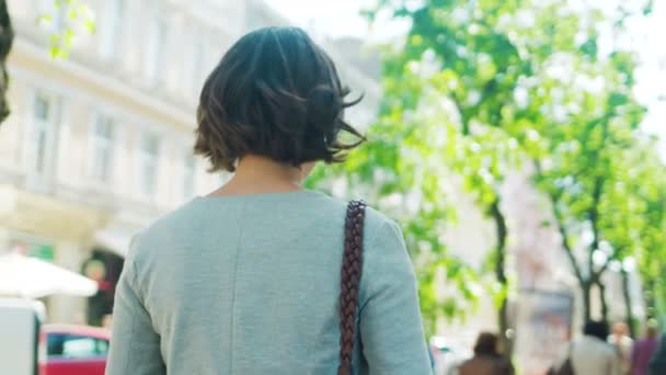 Hermosa joven mujer caminando, girando y sonriendo a la cámara en la calle de la ciudad. La cámara sigue detrás. Vista trasera — Vídeos de Stock