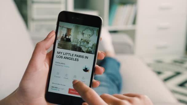 New York - 10 maart 2017: Vrouw handen zoeken kamers en hotels te huur op Airbnb app op smartphone liggend op de Bank. Close-up. Binnen schot. — Stockvideo