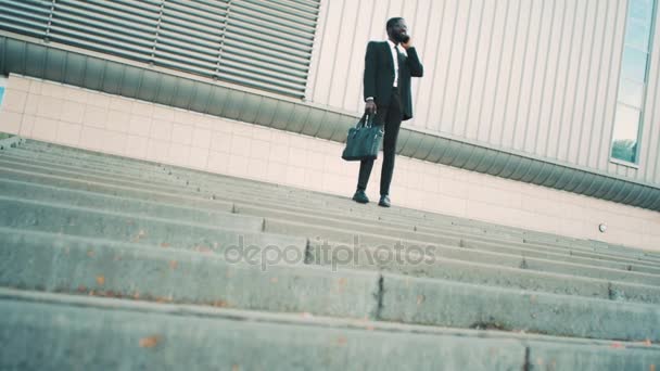 Atractivo hombre de negocios afroamericano balsck hablando por el teléfono celular, saludando de la mano a los colegios, mientras que de pie al aire libre en las escaleras cerca del gran centro de oficinas . — Vídeo de stock