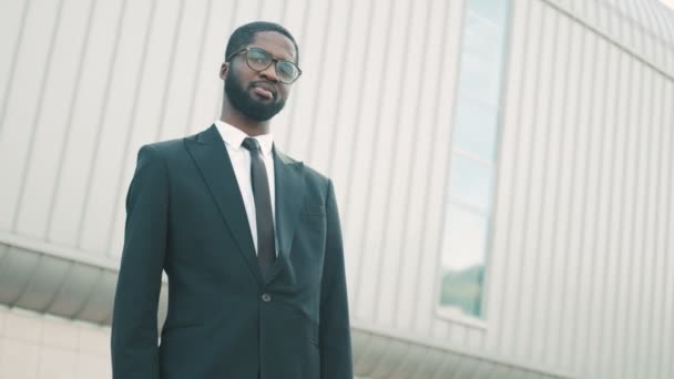 Joven hombre de negocios negro atractivo y seguro mirando a la cámara y sonriendo. De pie al aire libre cerca del gran edificio del centro de oficinas — Vídeos de Stock