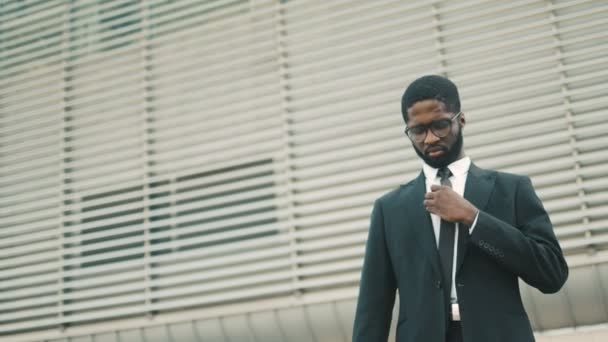 Joven y hermoso hombre de negocios afroamericano arreglando corbata y poniendo el teléfono celular en el bolsillo al aire libre cerca del centro de oficinas. Retrato plano — Vídeos de Stock