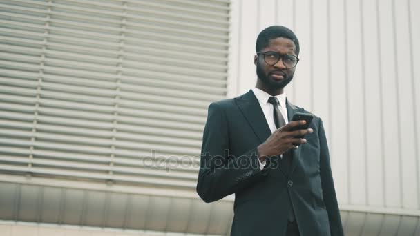 Молодой чернокожий американский бизнесмен пользуется смартфоном на открытом воздухе возле офисного центра. Бизнесмен читает новости, проверяет информацию, серфинг в Интернете по мобильному телефону — стоковое видео