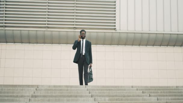 Atractivo joven hombre de negocios afroamericano hablando por teléfono celular mientras camina por las escaleras, pasos cerca del edificio del centro de oficinas . — Vídeo de stock