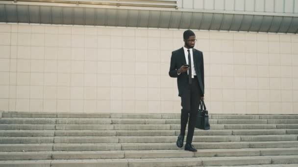 Attraktiva afrikanska amerikansk affärsman gick nedför trapporna, steg, med hjälp av smart telefon. Man läsa sms, surfa internet. Mannen kommer hem efter jobbet — Stockvideo