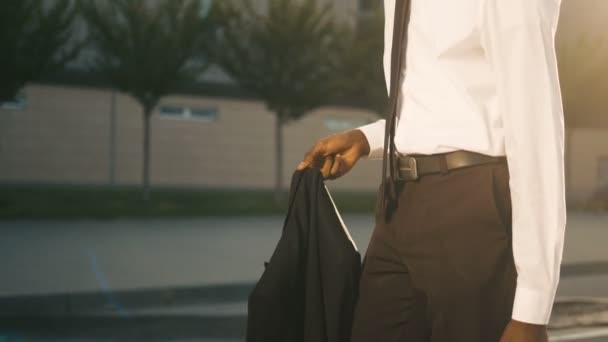 Atractivo hombre de negocios afroamericano negro caminando cerca del centro de oficinas sosteniendo la chaqueta y el bolso, sonriendo. El hombre se va a casa después del trabajo. Rayos solares. De cerca. — Vídeos de Stock