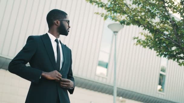 Feliz atraente empresário afro-americano em pé perto do prédio do centro de escritórios, olhando para os lados e direto para a câmera. Retrato tirado — Vídeo de Stock
