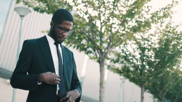 Confiant homme d'affaires afro-américain attrayant debout près du bâtiment du centre de bureaux, corrigeant veste et cravate et regardant la caméra. Portrait tourné — Video