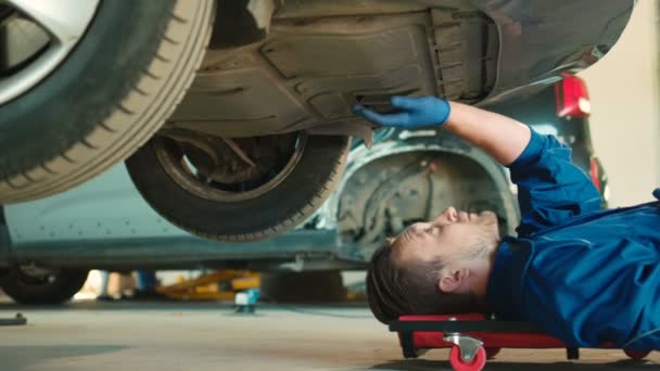 Kaukasischer junger Automechaniker, der den Zustand des auf dem Autoservice-Boden liegenden Fahrzeugs überprüft. Innenräume. — Stockvideo