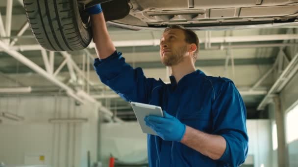 Giovane meccanico automobilistico concentrato caucasico mastica lo stato della ruota dell'auto e digita su tablet in autoservice. Interni . — Video Stock