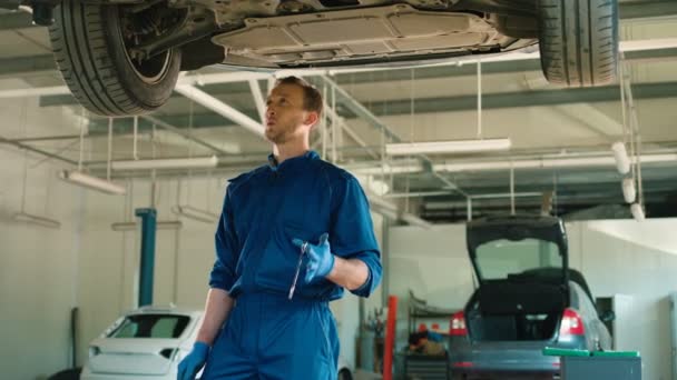 白种人的年轻汽车机械人穿着蓝色制服吹口哨和微笑。汽车修理工在维修服务使用扳手站。室内. — 图库视频影像