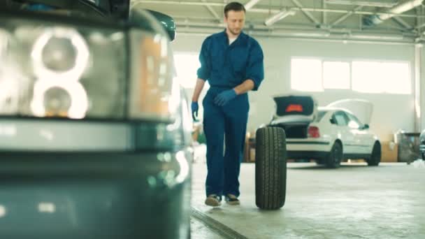 Kaukasiska unga stiliga bilmekaniker i blå uniform rullande hjul går på autoservice. Inomhus. Närbild. — Stockvideo
