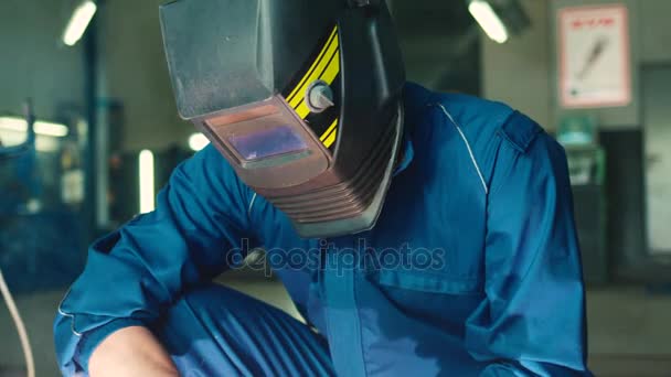 Портрет автомеханика в синей униформе и сварочном шлеме с помощью сварочного устройства на заднем плане автосервиса. В помещении . — стоковое видео