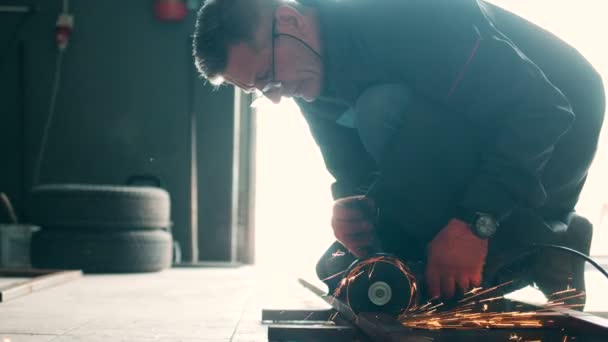 Ein mittelalter Automechaniker mit Brille sitzt beim Autoservice in blauer Uniform auf dem Boden und sägt den Eisenblock. Innenräume. — Stockvideo