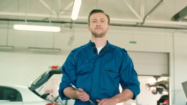 Kaukasische schöne Automechaniker Mann in blauer Uniform mit rauen Haaren hält einen Schraubenschlüssel in der Hand und komponiert Hände auf dem Autoservice Hintergrund. Innenräume. — Stockvideo