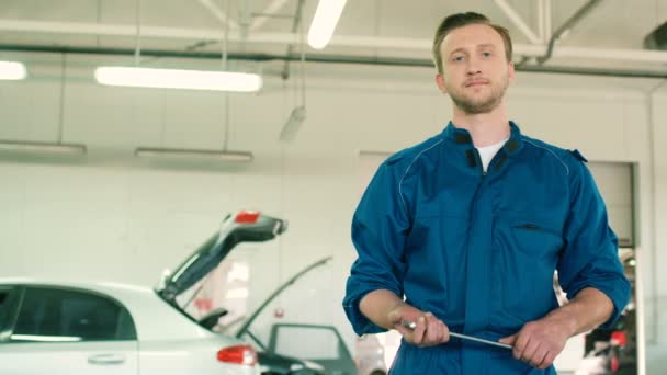 Hombre serio caucásico joven mecánico de automóviles en uniforme azul que compone las manos mientras sostiene una llave en la mano en el fondo de autoservicio. De interior . — Vídeos de Stock