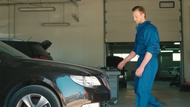 Schöner kaukasischer Automechaniker in blauer Uniform in Autoservice, der die Motorhaube des Autos hochzieht, um den Zustand der Mechanismen des Autos zu untersuchen. Innenräume. — Stockvideo
