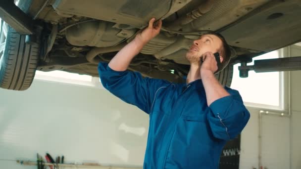 Kaukasischer junger Automechaniker in blauer Uniform, der mit einem Freund auf dem Handy telefoniert, während er sich den Zustand des Fahrzeugs ansieht. Innenräume. — Stockvideo