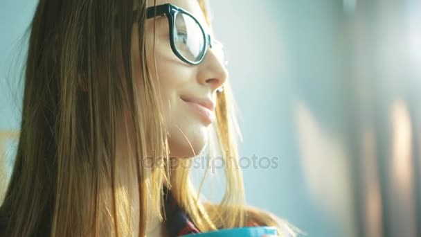 Retrato de cara de mujer cercana. Mujer con gafas, bebiendo café y mirando directamente a la cámara, sonriendo. Rayos solares. De cerca. — Vídeos de Stock