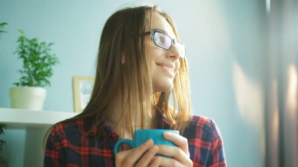 Jolie jeune femme portant des lunettes, buvant du café, du thé et regardant directement la caméra tout en étant assis sur le canapé à la maison. Prise de vue portrait. gros plan — Video