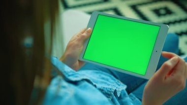 Beyaz tablet cihaz ile yeşil ekran kullanan kadın. Oturma odasında kanepede otururken sayfalarını kaydırma tablet, tutan kadın. Chroma anahtar. Omuz üzerinden görüntüleyin. Yakın çekim