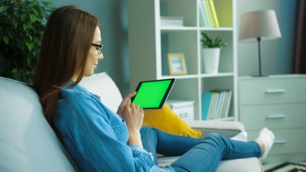 Mujer joven y relajada usando una tableta negra con pantalla verde mientras está sentada, acostada en el sofá de la sala de estar. Manos de mujer desplazándose, haciendo zoom de páginas. Clave de croma. vista lateral — Vídeos de Stock