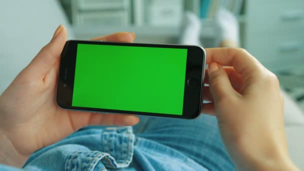 Mujer joven usando un teléfono inteligente negro con pantalla verde. Mujer sosteniendo el teléfono móvil en posición horizontal mientras está acostada en el sofá de la sala de estar. Clave de croma. Vista sobre el hombro. De cerca. — Vídeo de stock