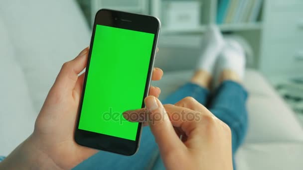Young woman using black smart phone with green screen. Woman holding mobile phone in vertical position while lying on the couch in the living room. Chroma key. Close up. Top view — Stock Video